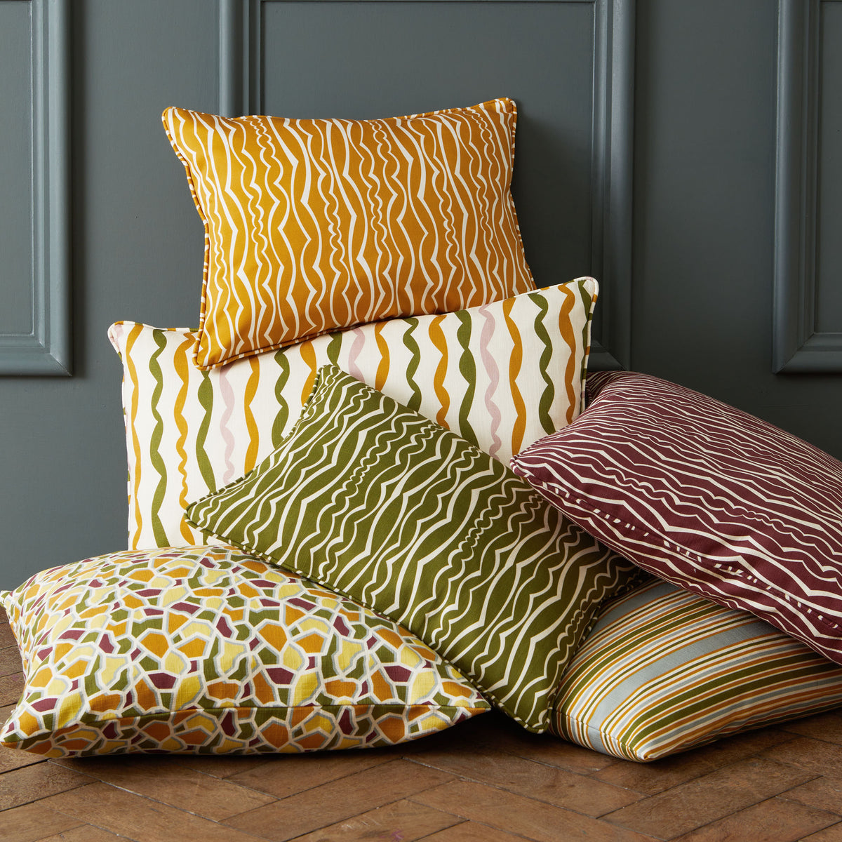 A collection of six decorative throw pillows with various patterns and colors, stacked on a wooden floor against a dark gray paneled wall.