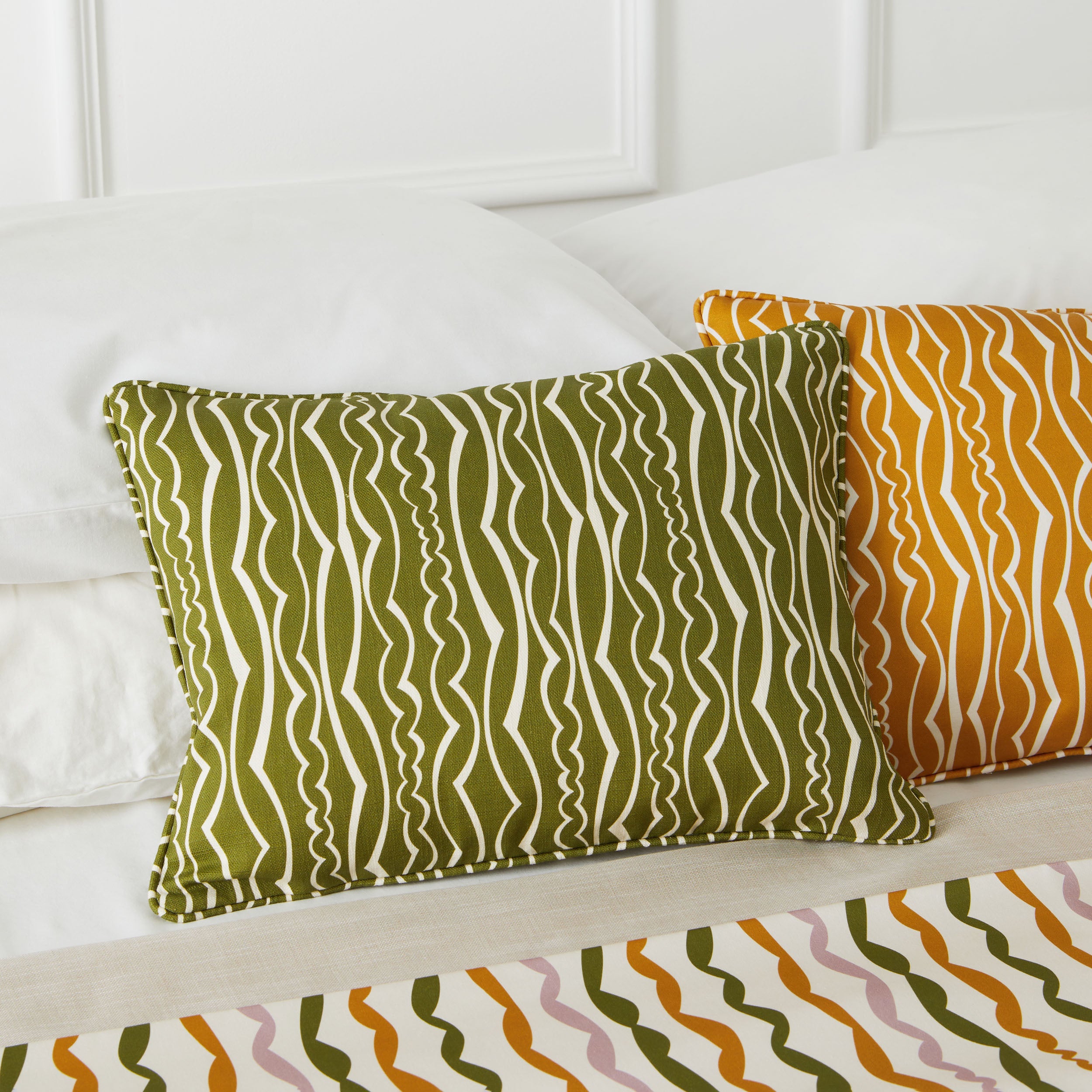 Two decorative throw pillows on a bed: one olive green with white wavy vertical stripes and one mustard yellow with orange wavy vertical stripes, with a matching patterned bedspread visible at the bottom.