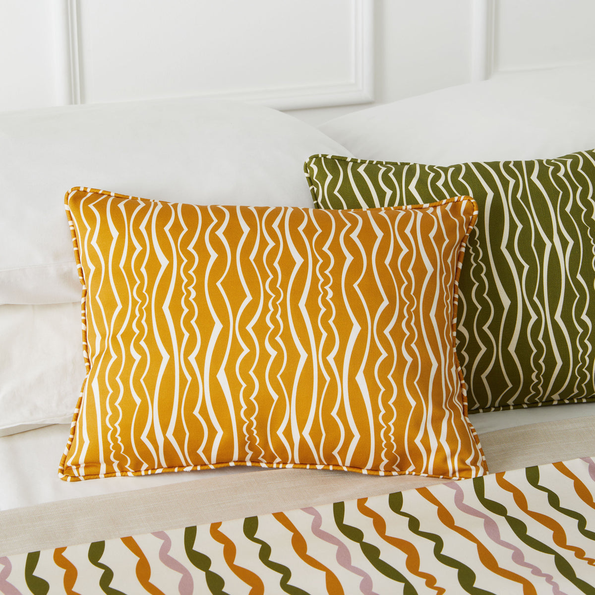 Two decorative throw pillows on a bed: one in mustard yellow with white wavy vertical stripes, and one in olive green with white wavy vertical stripes. A matching patterned bedspread is visible at the bottom.