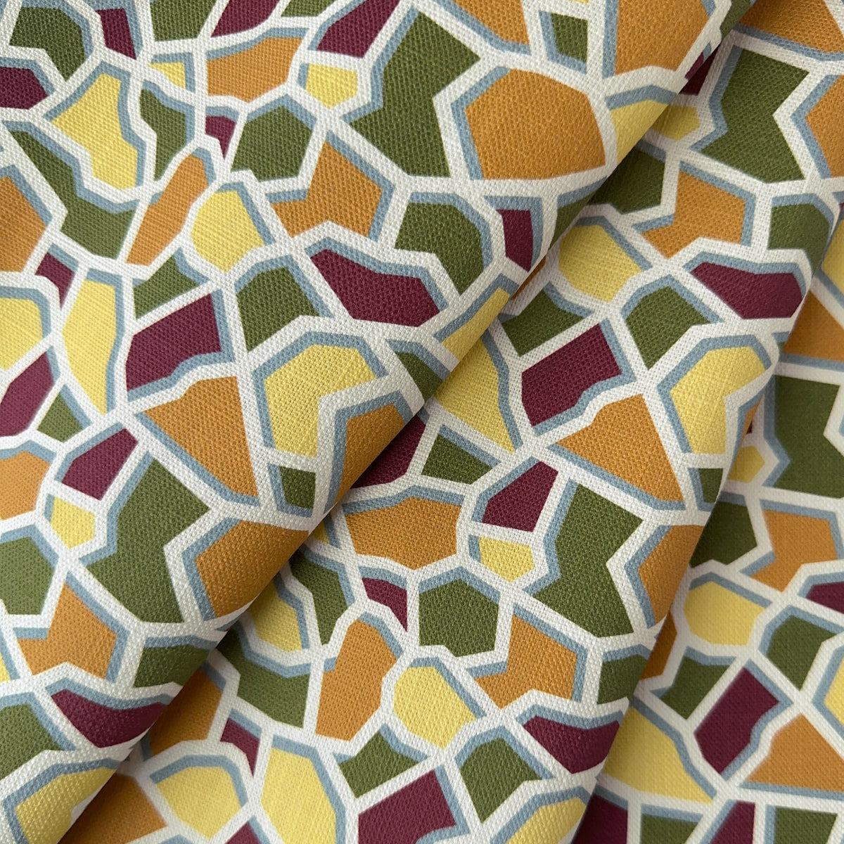 A close-up of a printed linen fabric with an abstract mosaic pattern of irregular polygon shapes in yellow, orange, olive green, burgundy, and cream.