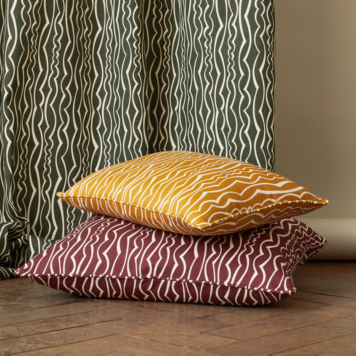 Two decorative throw pillows stacked on a wooden floor in front of a green patterned curtain. The top pillow is golden yellow with an ivory wavy stripe pattern, and the bottom pillow is burgundy with a matching ivory wavy stripe pattern.