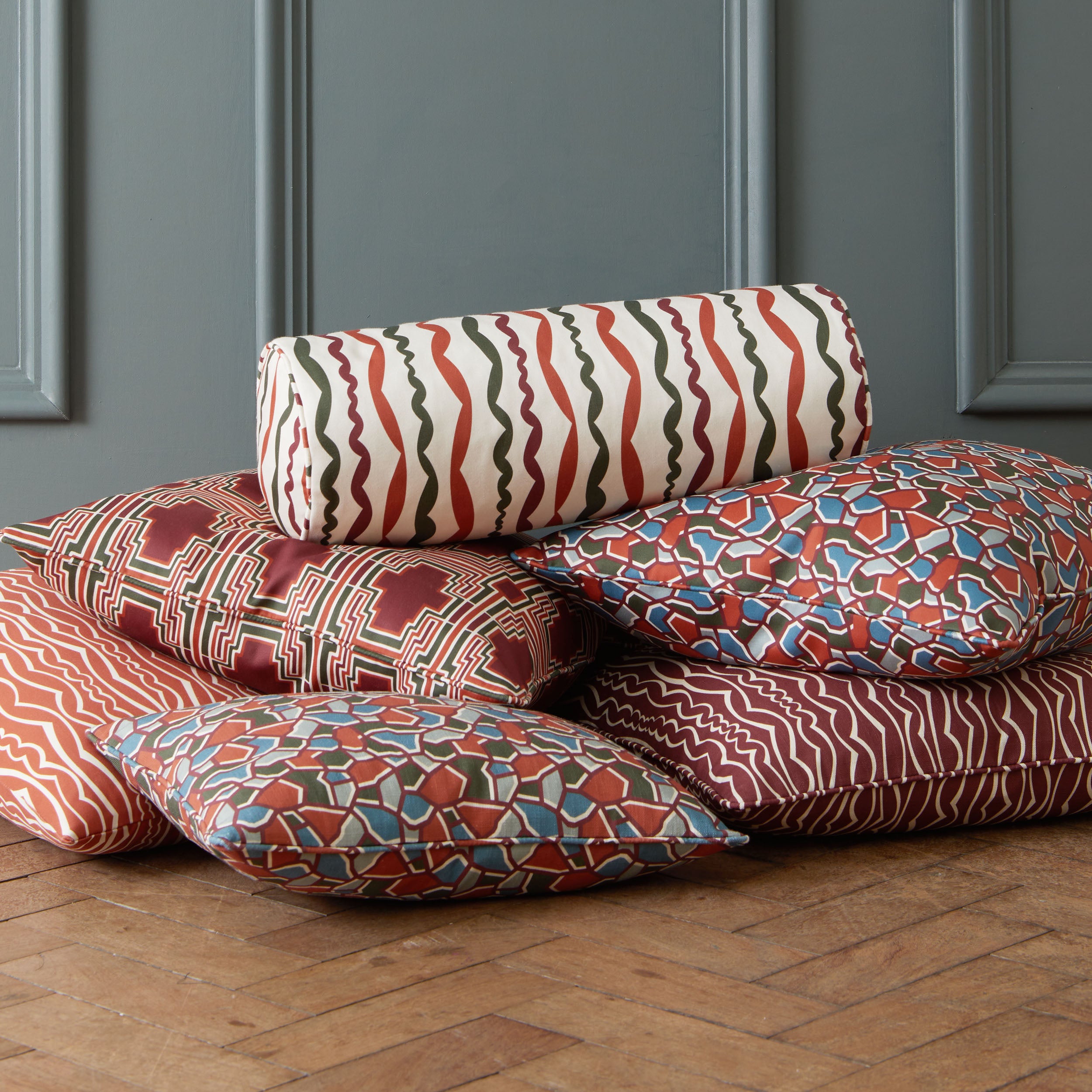 Cushion stack of 6 cushions in a variety of geometric designs, sitting on a wooden floor against a blue grey wall