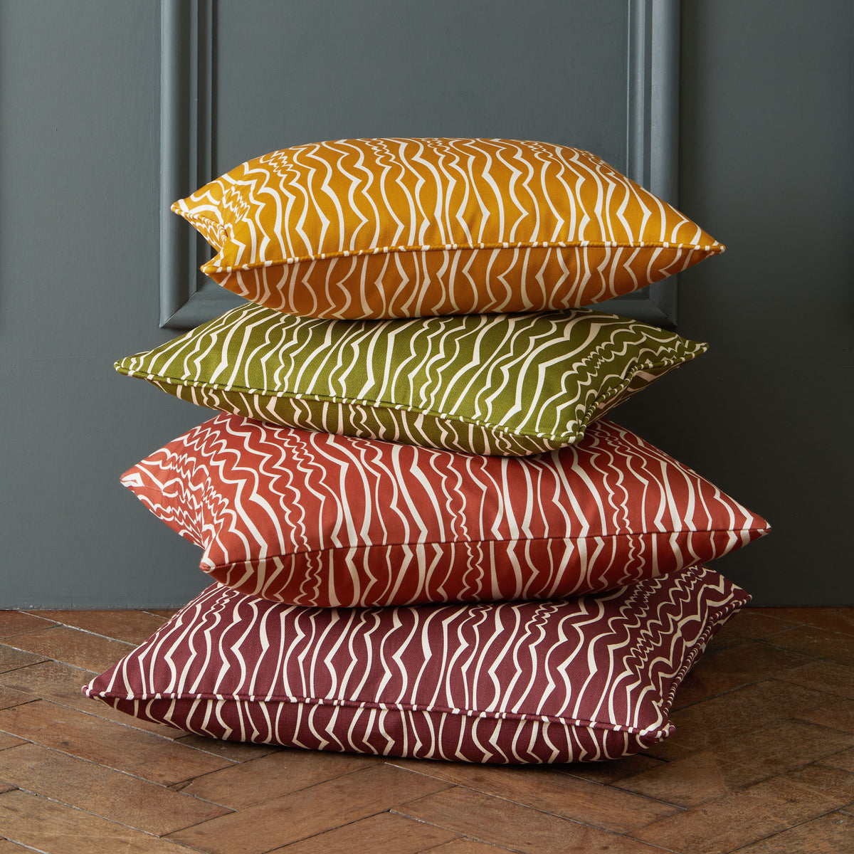 Stack of 4 colourful printed cushions featuring ivory stripes, against a grey wall.