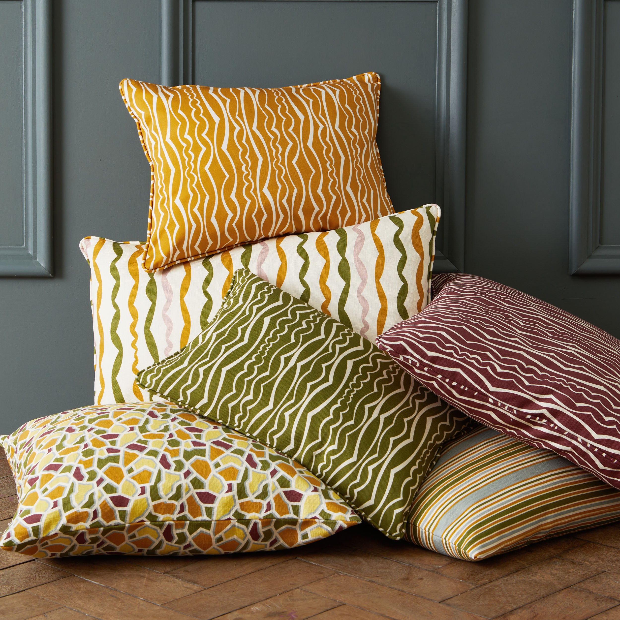 A collection of six decorative throw pillows with various patterns and colors, stacked on a wooden floor against a dark gray paneled wall.