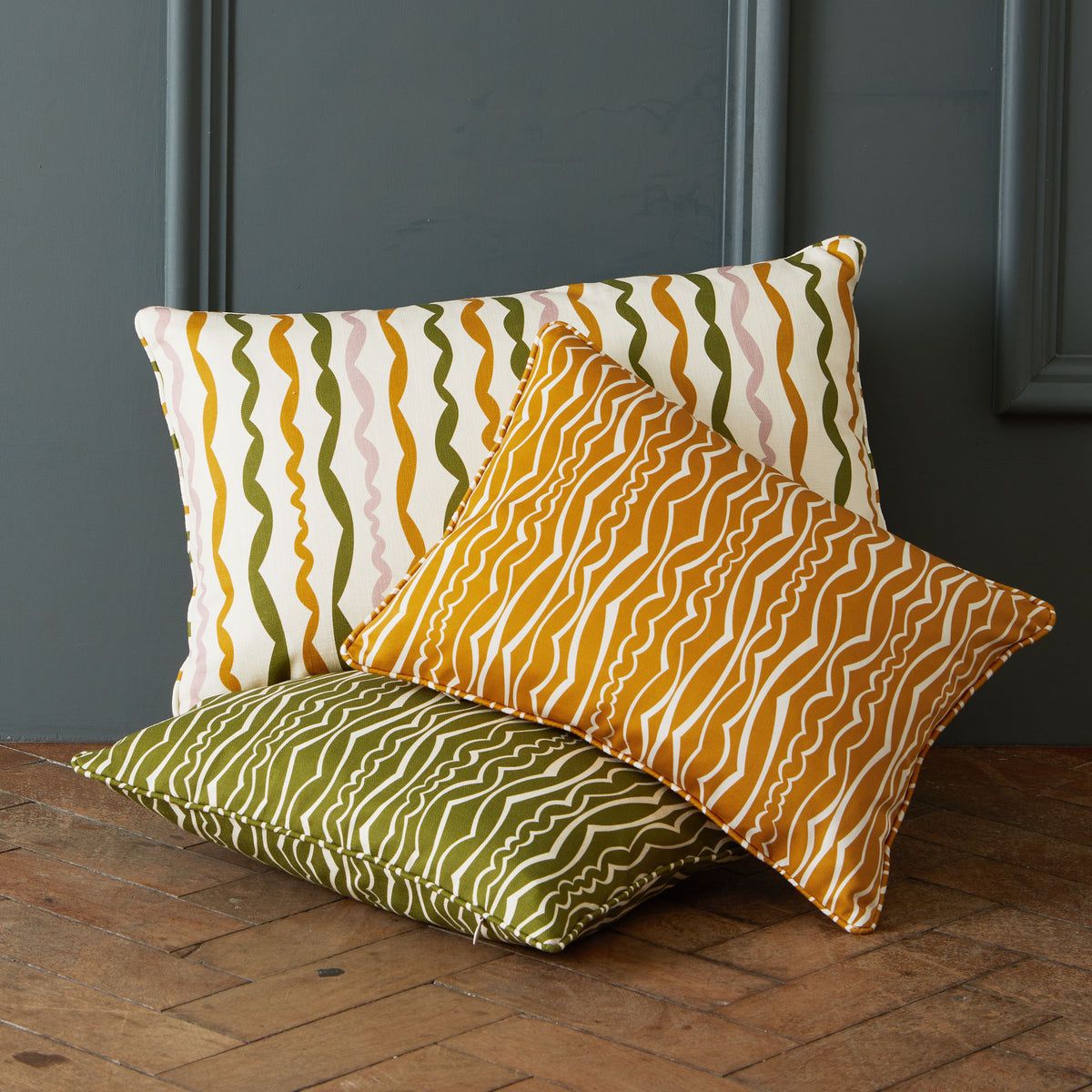 Three decorative throw pillows with wavy stripe patterns in yellow, green, pink, and cream, arranged on a wooden floor against a dark gray paneled wall.