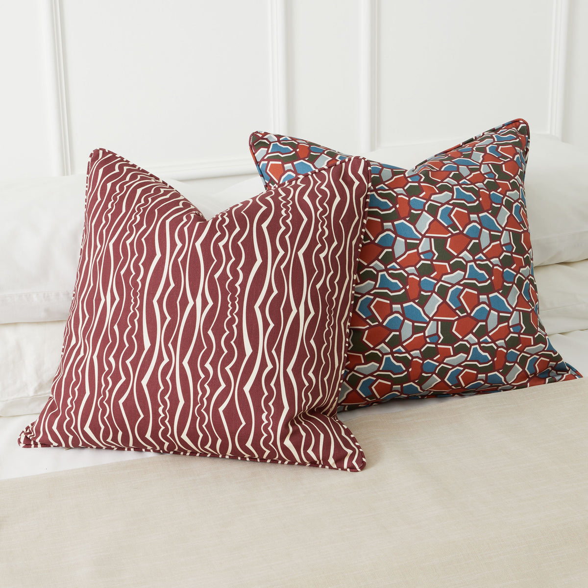 Two decorative throw pillows on a bed. The front pillow has a burgundy and ivory wavy stripe pattern, and the back pillow has a multicolor mosaic tile pattern in burgundy, blue, and forest green.
