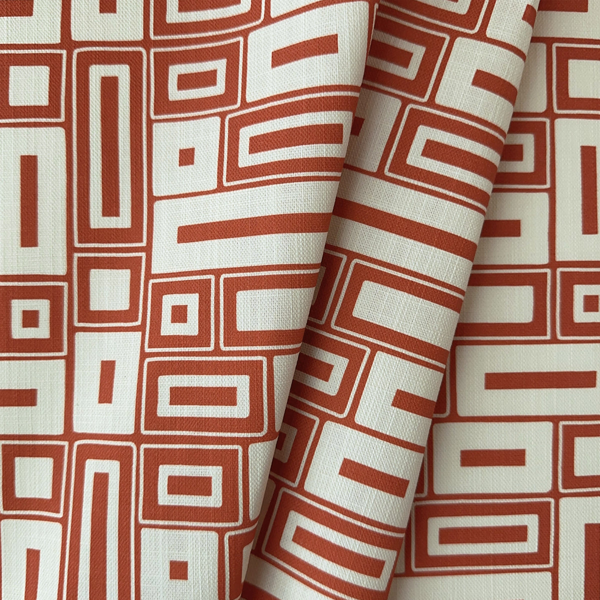 A close-up of patterned fabric with a terracotta red and off-white geometric design, featuring a central fold that highlights the textile's texture.