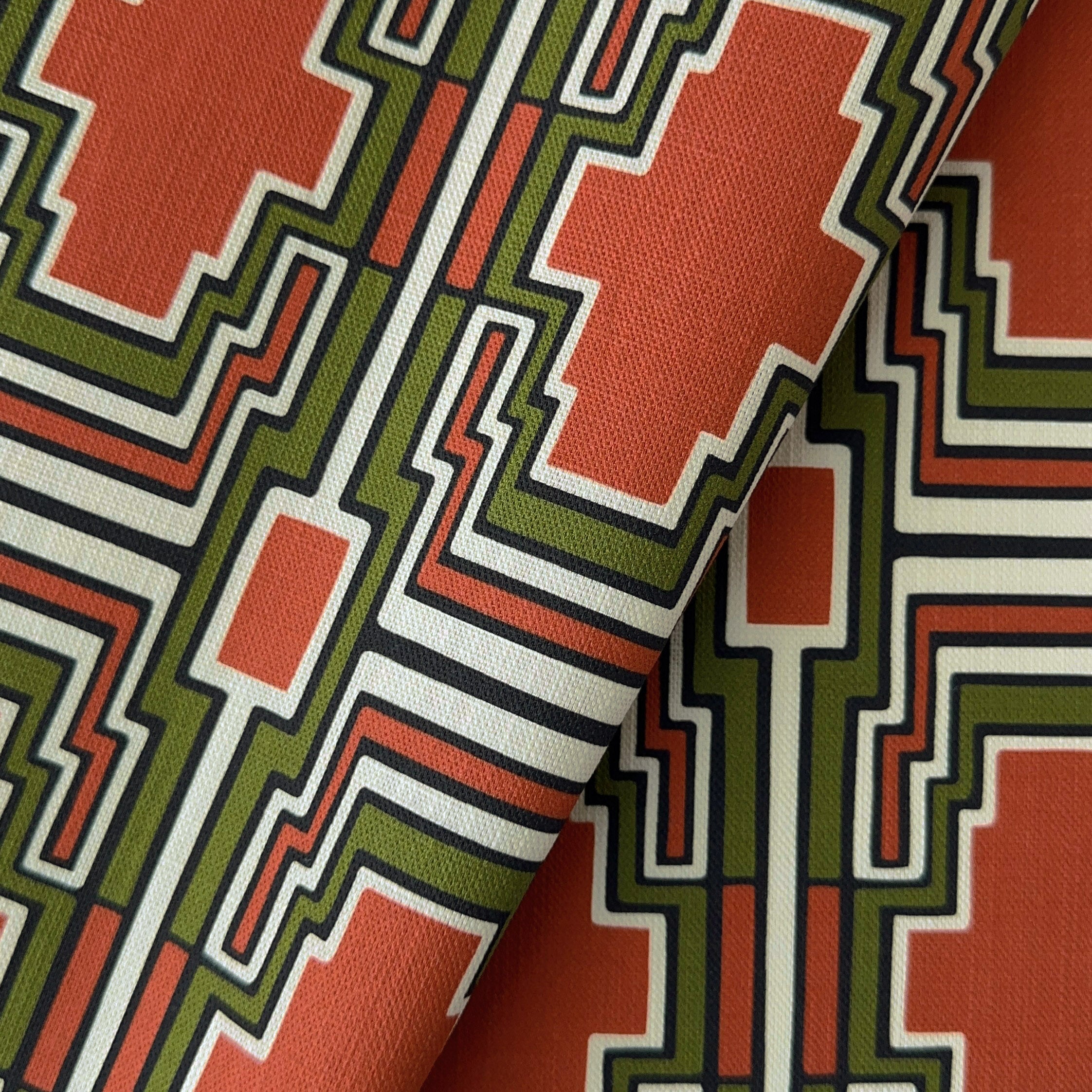 A close-up of a folded textile with a bold geometric pattern in terracotta orange, olive green, black, and off-white.