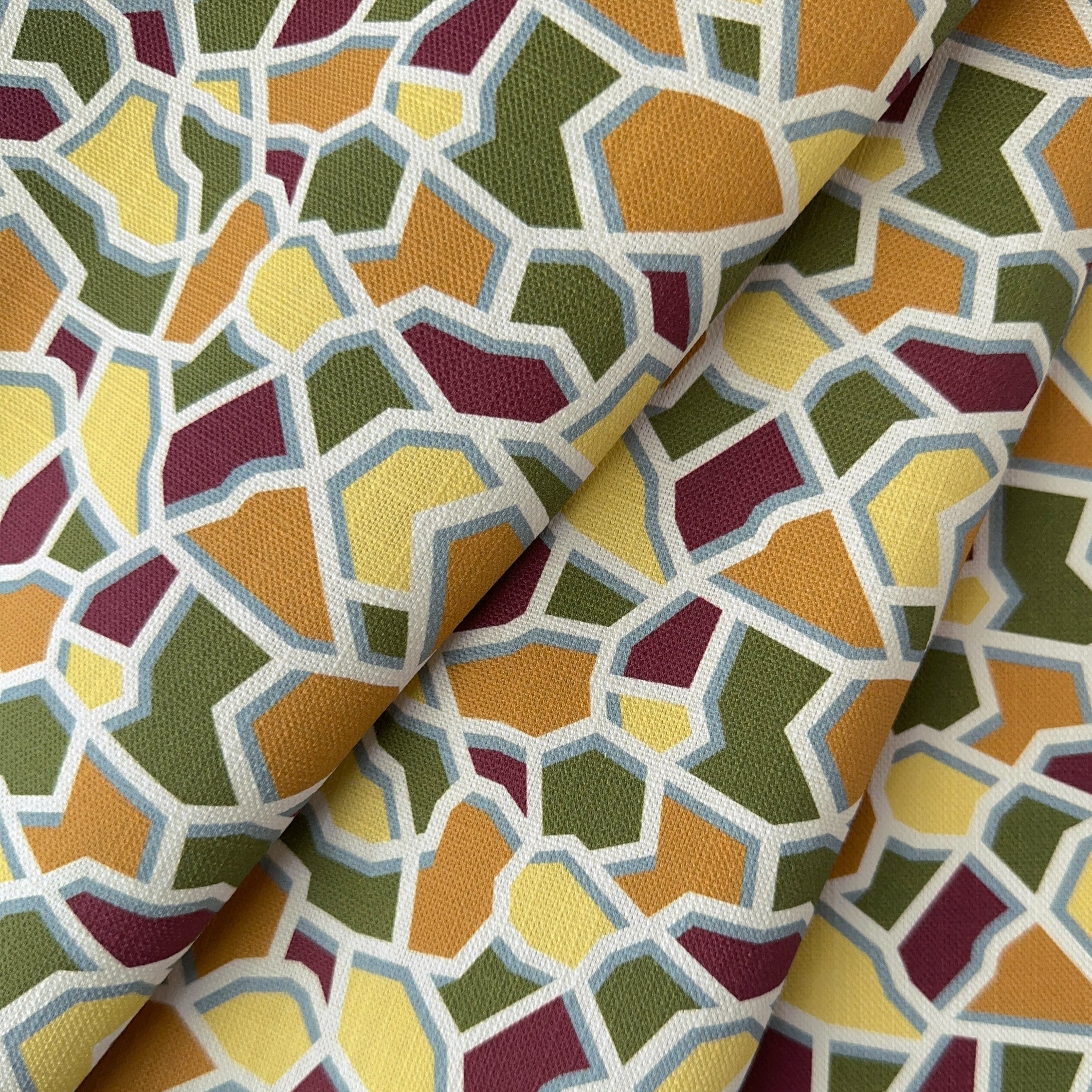 A close-up of a printed linen fabric with an abstract mosaic pattern of irregular polygon shapes in yellow, orange, olive green, burgundy, and cream.