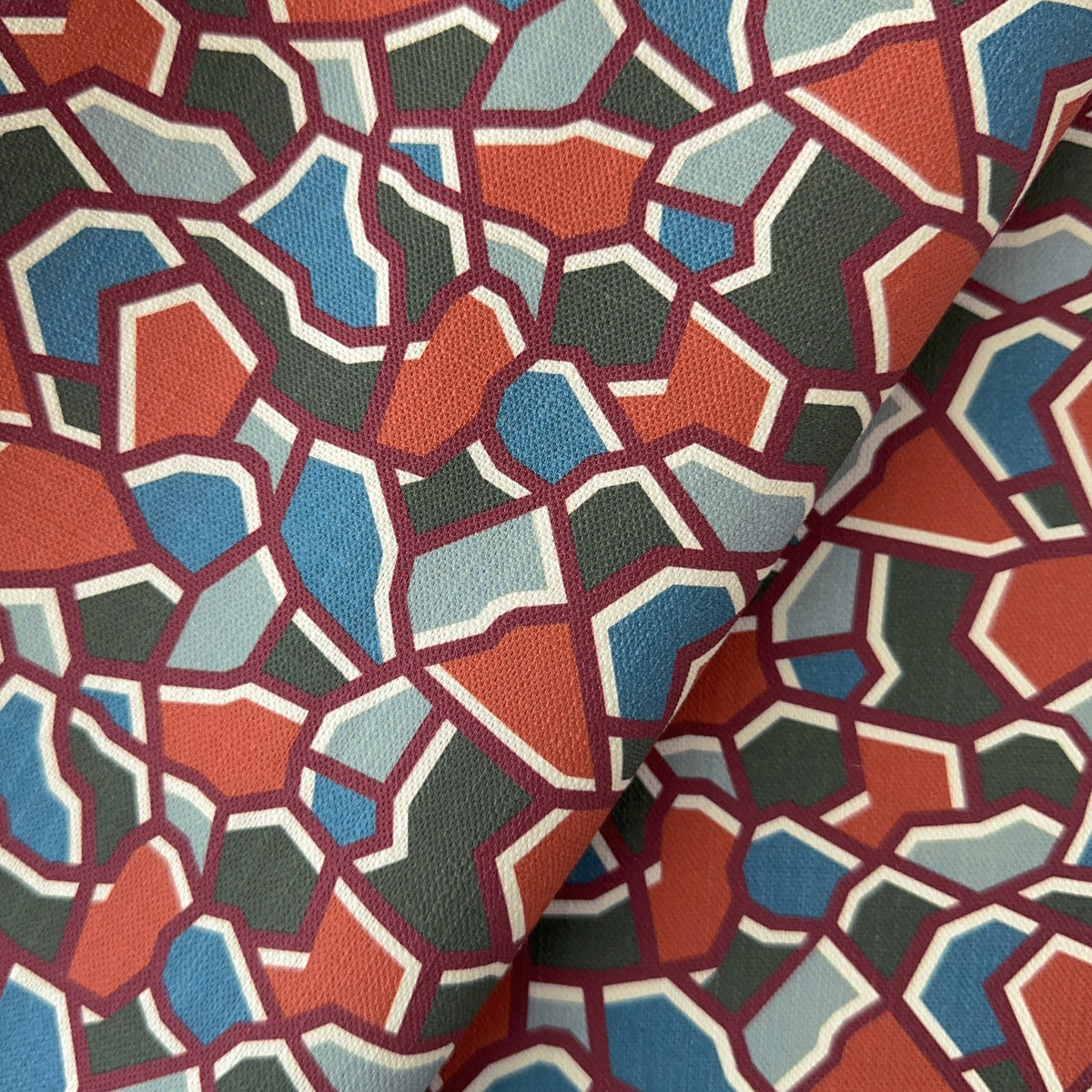 A close-up of patterned fabric with an abstract geometric mosaic design in terracotta, slate blue, dark green, light blue, cream, and burgundy.